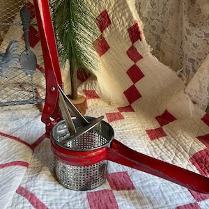 May include: A vintage red potato ricer with a stainless steel perforated bowl and long handle. The kitchen tool is designed for mashing potatoes and other soft foods. The ricer is placed on a white quilt with red diamond patterns.