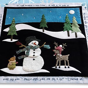 May include: A winter-themed felt art piece featuring a snowman, a reindeer, and a small animal. The snowman wears a hat, scarf, and mittens. The reindeer has a hat. The background depicts a snowy landscape with trees and a night sky. Text is included.