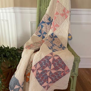May include: A vintage quilt draped over a green wicker chair. The quilt features a cream-colored background with patches of blue, pink, and patterned fabrics. The quilt's design includes pinwheel and diamond shapes.