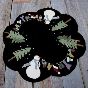 May include: A black, scalloped-edge placemat with a winter theme. It features two snowmen, green pine trees, and a string of hanging winter accessories. The design is set against a black background with white snowflake accents.