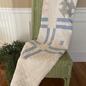 May include: A white quilted blanket featuring a geometric pattern with light blue stripes and grey and red plaid accents. The fabric has a diamond-stitched texture throughout, with a rectangular shape draped over a light green wicker chair.