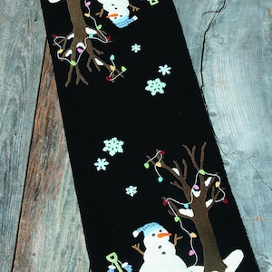 May include: Black felt table runner with a winter scene. Features two snowmen, trees with string lights, and snowflakes. One snowman wears a blue hat and scarf. The other snowman is upside down. A red sled is also present.