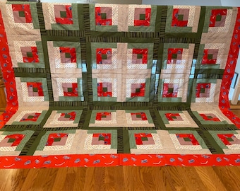 Könnte beinhalten: Eine handgefertigte Steppdecke mit Patchwork-Design. Die Steppdecke weist ein sich wiederholendes Muster aus roten, grünen, weißen und beigen Quadraten auf. Der Rand ist rot mit einem blau-weißen Muster. Die Steppdecke wird auf einem Holzboden ausgestellt.
