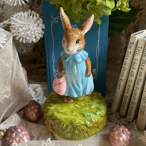 May include: A ceramic figurine of Peter Rabbit, wearing a blue jacket and holding a pink purse, stands on a green grassy base. The figurine is set against a backdrop of books and decorative elements, including lace and faux flowers.