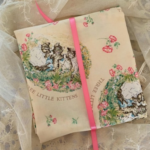 May include: A folded piece of cream-colored fabric featuring a repeating illustration of three kittens in a garden with pink flowers and green foliage. The text "THREE LITTLE KITTENS" is printed on the fabric, which is secured by a bright pink ribbon.