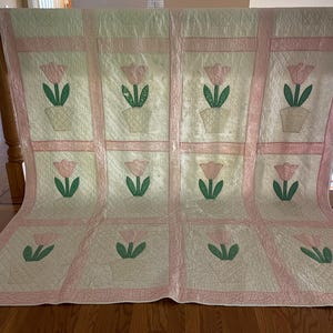 May include: A rectangular quilt featuring a grid of twelve blocks, each displaying a pink tulip in a cream-colored pot with green leaves. The blocks are separated by light pink sashing, and the entire quilt has a cream-colored background.