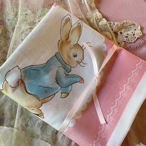 May include: A folded white and pink fabric item featuring a Peter Rabbit illustration in a blue jacket. The item is tied with a pink ribbon. The border is pink with a white scalloped edge and a white floral design. The background includes lace and a pink placemat.