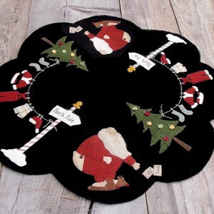 May include: Black Christmas-themed placemat with a scalloped edge. The design features Santa Claus, Christmas trees, and a North Pole sign. Red, green, white, and tan accents create a festive holiday scene. The placemat is likely made of felt or a similar material.