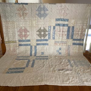 May include: A rectangular quilt featuring a white base fabric with a repeating geometric pattern in muted shades of blue, pink, and grey. The design consists of pieced blocks with cross and square motifs, finished with visible hand-stitched quilting.