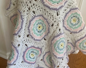 Könnte beinhalten: Eine handgefertigte, weiße Häkeldecke mit einem sich wiederholenden kreisförmigen Muster. Jeder Kreis zeigt Pastellfarben: Rosa, Blau, Grün und Gelb. Die Decke ist über einen kleinen Hocker drapiert, um ihre Textur und ihr Design zu zeigen. Ein gemütlicher, dekorativer Artikel.