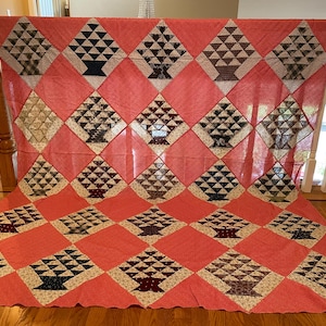 May include: A large rectangular quilt featuring a repeating pattern of diamond-shaped blocks. Each block contains a pieced basket motif in various dark colors and patterns, set against a vibrant coral pink background fabric throughout the quilt.