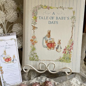 May include: A vintage-style baby book titled "A Tale of Baby's Days" with illustrations of rabbits and flowers. The book is displayed on a white metal stand, accompanied by a matching bookplate and three pink glass ornaments.