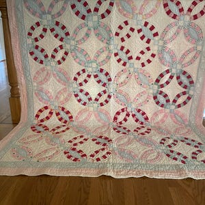 May include: A double wedding ring quilt featuring a cream-colored background with interlocking rings in shades of red, pink, and light blue. The fabric displays various floral and geometric patterns, finished with a light pink border and stitching.