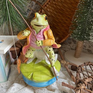 May include: A ceramic frog figurine dressed in a pink shirt and yellow coat, holding a fishing rod with a carrot lure. The frog is perched on a green lily pad with a blue base. The figurine has green spots and is part of a whimsical collection.