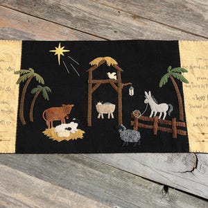May include: A black felt nativity scene with a star, palm trees, and various animals. The scene includes a cow, sheep, donkey, and a manger. Text is on the left and right sides of the scene.