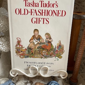 May include: A book titled "Tasha Tudor's Old-Fashioned Gifts" is displayed on a white metal stand. The book cover features an illustration of people crafting. Sewing supplies, including thread and scissors, are visible in the foreground.