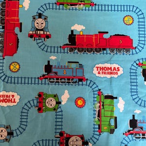 May include: A light blue fabric featuring colorful illustrations of Thomas the Tank Engine and his friends. The design includes various train engines, train tracks, and the text "Thomas & Friends" in white clouds.