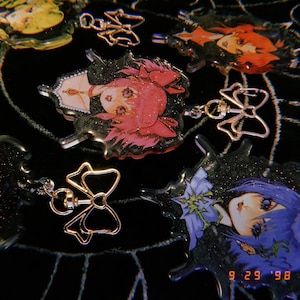 May include: A collection of six colorful anime character charms with glitter accents. Each charm features a different character with a unique hairstyle and expression. The charms are attached to gold-colored metal rings with bows.