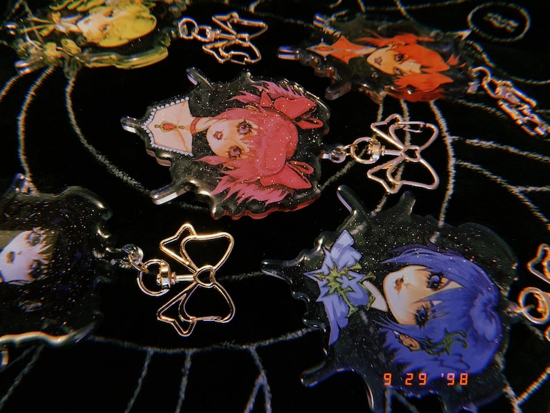 May include: A collection of six colorful anime character charms with glitter accents. Each charm features a different character with a unique hairstyle and expression. The charms are attached to gold-colored metal rings with bows.