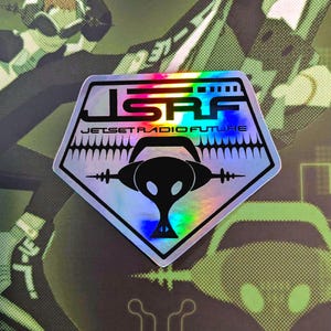 Op de afbeelding: Holografische sticker met de tekst "JSRF JET SET RADIO FUTURE" in zwart en wit. De sticker heeft een vijfhoekige vorm met een zwarte koptelefoongrafiek in het midden. De achtergrond is een camouflagepatroon.