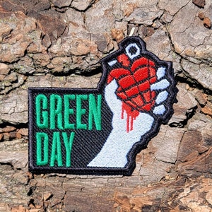 May include: Green Day band patch with a green and black background and the band's name in green text. The patch features a white hand holding a red and black heart-shaped grenade with a white fuse.