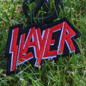 May include: A black and red embroidered patch with the word "Slayer" in white lettering. The patch is made of fabric and has a rough texture.