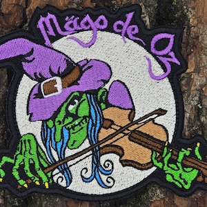 May include: Embroidered patch featuring a green witch with purple hat and long blue hair playing a violin. The witch is in front of a white moon with the text "Mago de la" in purple.