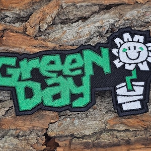 May include: Green Day band logo embroidered patch with a green and black color scheme. The logo features the band name "Green Day" in a stylized font. A white flower with a smiley face is on the right side of the patch.
