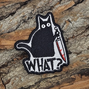 May include: Black cat patch with a white outline, holding a bloody knife. The word "WHAT?" is written in white, outlined in black. The patch is on a textured, brown surface.