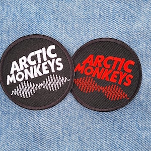 May include: Two black and white embroidered patches with the text "Arctic Monkeys" in white and red lettering. The patches feature a stylized sound wave design.
