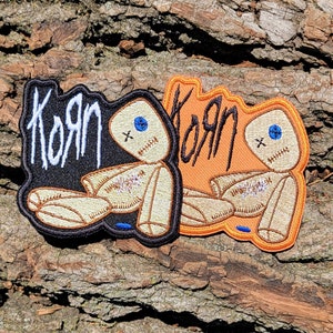 May include: Two embroidered patches featuring a ragdoll design with the word "Korn" in white lettering. The patch on the left is black with a tan ragdoll, and the patch on the right is orange with a tan ragdoll.