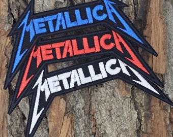 May include: Three embroidered patches with the Metallica logo in blue, red, and white. The patches are layered on top of each other, with the blue patch on top.