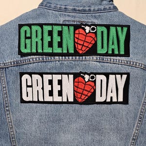 May include: A light blue denim jacket with two patches. The patches feature the words "GREEN DAY" in green and white, with a red heart-shaped grenade graphic. The patches are rectangular and sewn onto the back of the jacket.