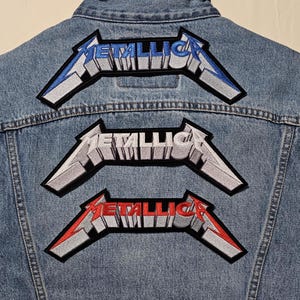 May include: A light blue denim vest with three embroidered "Metallica" patches. The patches feature the band's logo in white, red, and blue, with a black border. The vest has a classic design with a collar and button-up front.