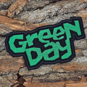 May include: Green Day embroidered patch with black outline and green lettering. The patch is on a brown wooden background.