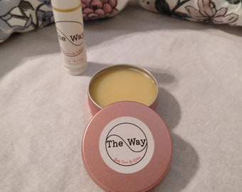 May include: A pink tin with a white label that says "The Way Body Care by Kiera" and a yellow lip balm in a white tube with a pink label that says "The Way Lip Balm".