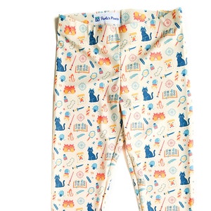 May include: Cream-colored leggings with a whimsical pattern featuring blue cats, orange flames, and other magical symbols. The leggings have a comfortable, stretchy fit.