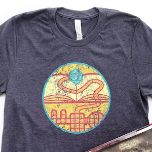 May include: A dark heather grey t-shirt with a vintage-style graphic of a city skyline and a blue and yellow dice. The graphic is surrounded by a red circle with a yellow border. The t-shirt is lying on a white surface next to an open book with a map of a dungeon.
