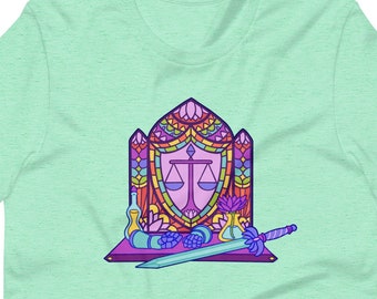 May include: A light green t-shirt featuring a colorful graphic. The graphic depicts a stained-glass window design with scales, a sword, and other elements. The design is outlined in black and filled with shades of purple, orange, and blue.