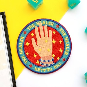 May include: A red, blue, and yellow embroidered patch with a hand with glowing stars and the text "THE HEALER" repeated around the edge. The patch is surrounded by turquoise blue dice with gold numbers.