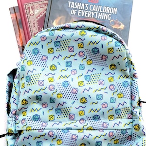 May include: A light blue backpack with a geometric pattern of dice, squares, and wavy lines. The backpack has black straps and a zipper closure. The backpack is filled with two books, one red and one blue, with the text "Tasha's Cauldron of Everything" visible on the blue book.