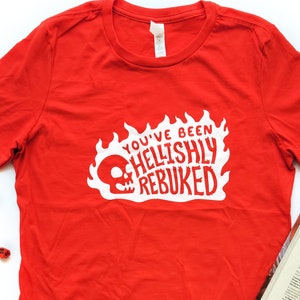 May include: Red t-shirt with a white graphic that says "You've Been Hellishly Rebuked" with a skull and flames.
