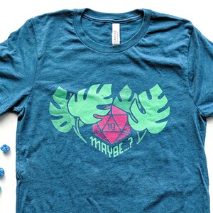 Puede incluir: Camiseta azul turquesa con un dise&ntilde;o de hojas de monstera verdes y un dado rojo de 10 caras con el texto "MAYBE...?"