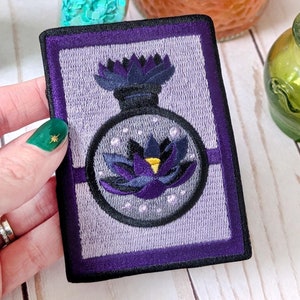 May include: Purple embroidered patch with a black and white potion bottle with a flower inside. The bottle has a purple lid and a black flower with a yellow center.