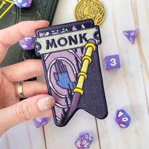 May include: Purple and black embroidered patch with a hand reaching out from a circle and a staff with a yellow and white design. The patch has the word "MONK" written on it.