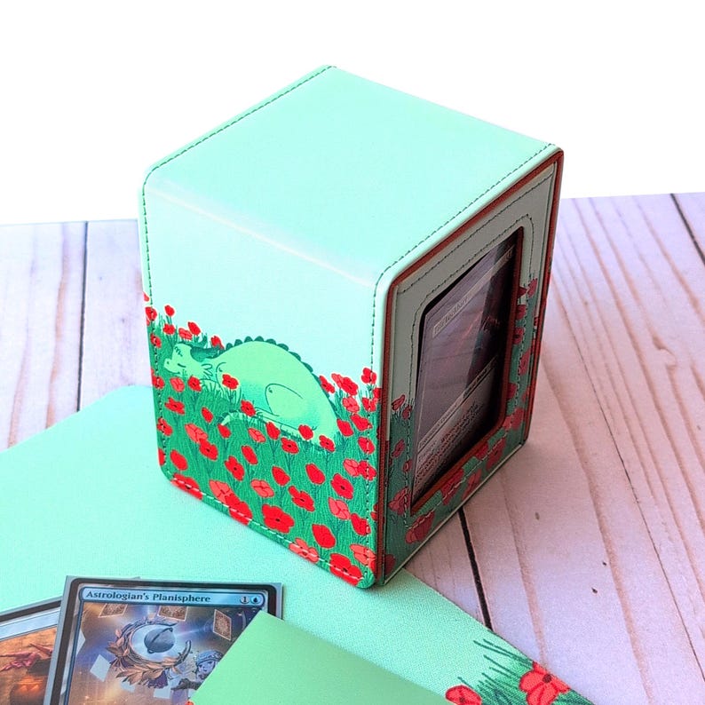 Puede incluir: Caja de almacenamiento de tarjetas verde menta con ventana ribeteada en rojo. La caja presenta un dise&ntilde;o caprichoso de un drag&oacute;n verde descansando en un campo de amapolas rojas. La caja es cuadrada y tiene un interior verde menta a juego.