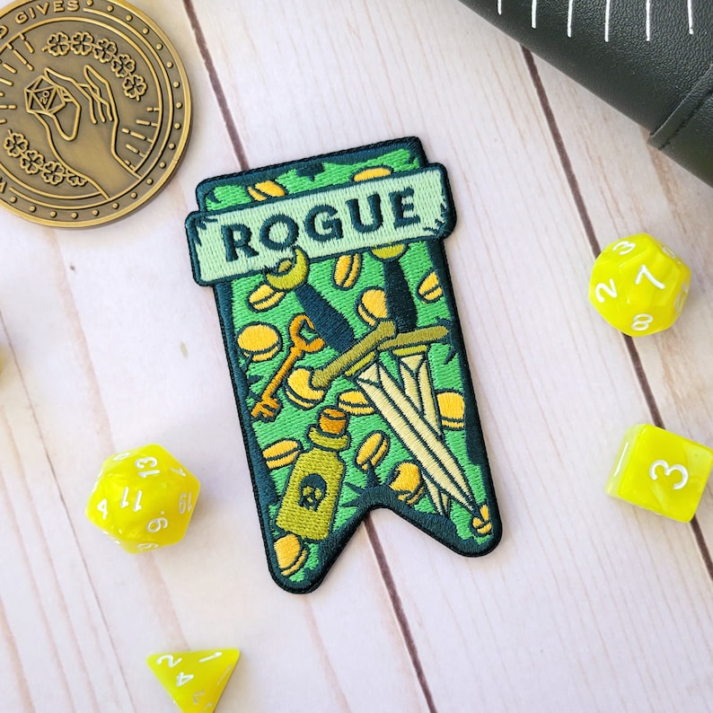 Pu&ograve; includere: Toppa di feltro verde con la parola "ROGUE" in testo bianco. La toppa presenta uno sfondo verde con un pugnale giallo, una chiave e una boccetta di pozione con un teschio e ossa incrociate. La toppa &egrave; circondata da dadi gialli.