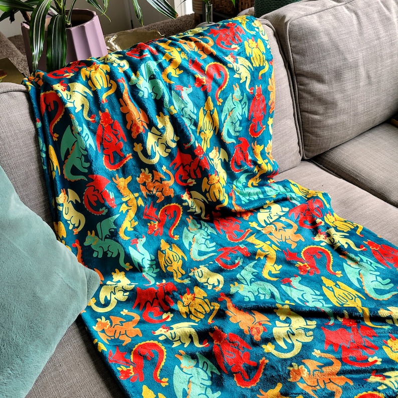 Pu&ograve; includere: Una coperta turchese con un motivo ripetuto di draghi colorati in rosso, giallo, arancione e verde. La coperta &egrave; drappeggiata su un divano grigio, creando un'atmosfera accogliente. La coperta &egrave; realizzata in un materiale morbido.