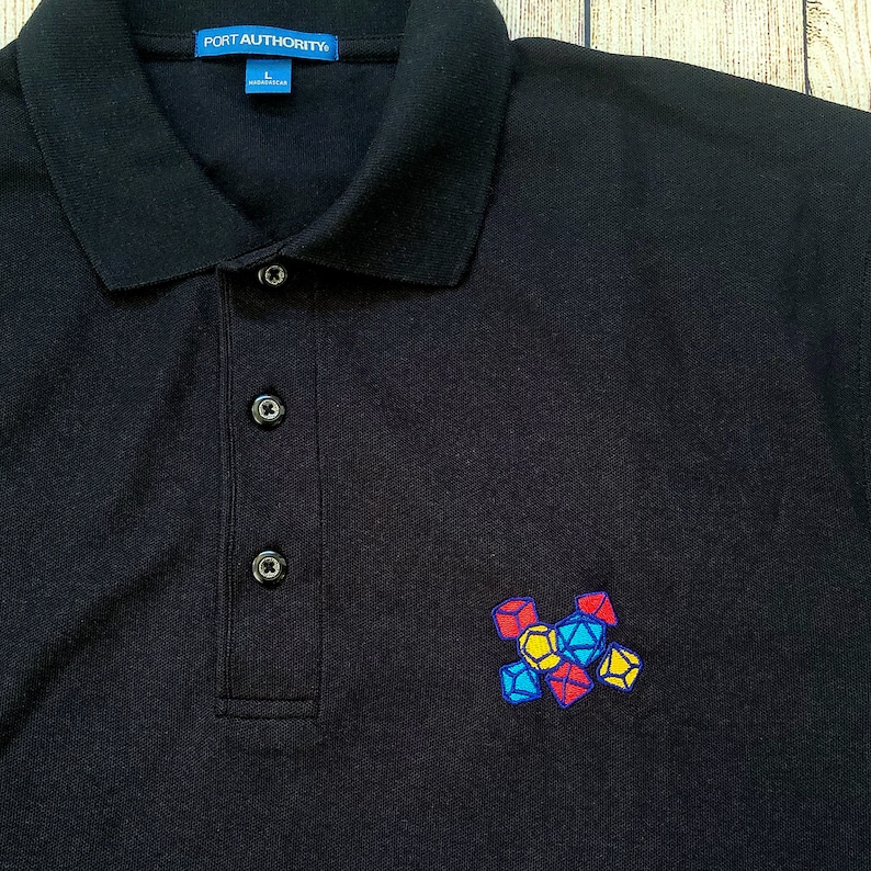 May include: Black polo shirt with a small embroidered design of colorful dice on the right side of the shirt. The shirt has a blue label with the text "PORT AUTHORITY" and the size "L" on the left side of the collar.