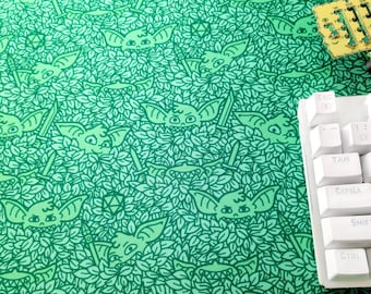 Puede incluir: Alfombrilla de ratón verde con un patrón repetido de murciélagos de dibujos animados, espadas y dados. El diseño está sobre un fondo de hojas. Un teclado blanco es parcialmente visible, junto con una pequeña placa de circuito.